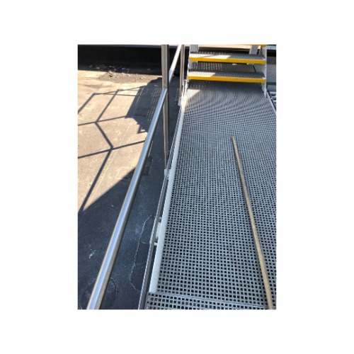 Metalowa konstrukcja schodów z podestem i poręczą, z żółtymi elementami antypoślizgowymi na stopniach, widoczna z boku w silnym oświetleniu słonecznym.