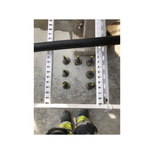 Metalowe kotwy w betonie, zabezpieczone żółtymi zaślepkami, przymocowane do białej konstrukcji. Widoczne buty robocze w dolnej części kadru.