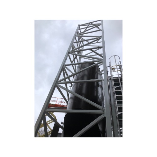 Wysoka konstrukcja stalowa z czarnym zbiornikiem w tle, widok z dołu pod kątem. Szara, pochmurna pogoda. Elementy konstrukcji spawane.