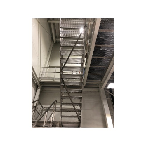 Metalowe schody z ażurowymi stopniami w jasnym wnętrzu hali. Widok z dołu na konstrukcję schodów i platformy. Elementy spawane i skręcane.