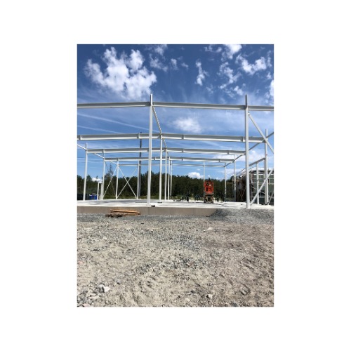 Stalowa konstrukcja szkieletowa budynku w trakcie montażu, widoczny podest i niebo. W tle drzewa i sprzęt budowlany. Elementy spawane na miejscu.