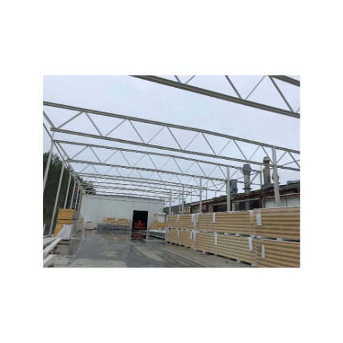 Konstrukcja stalowa hali w trakcie budowy, widoczne elementy spawane, płyty warstwowe, szkielet dachu i fragment budynku w tle.