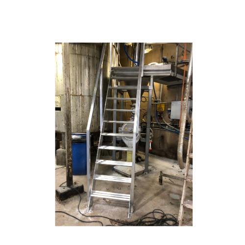 Metalowe schody przemysłowe z platformą w hali produkcyjnej, widoczne rury i zbiornik. Konstrukcja spawana, surowe wykończenie, oświetlenie halogenowe.