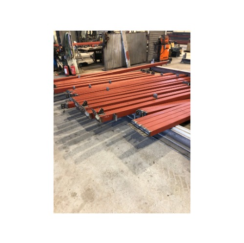 Czerwone, pomalowane profile stalowe, przygotowane do spawania, leżące na szarej posadzce warsztatu. W tle widoczny sprzęt spawalniczy.