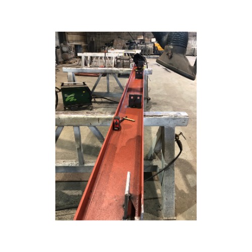 Czerwona belka stalowa z przyspawanymi elementami, leżąca na metalowych kozłach w warsztacie. Widoczny sprzęt spawalniczy i odciąg oparów.