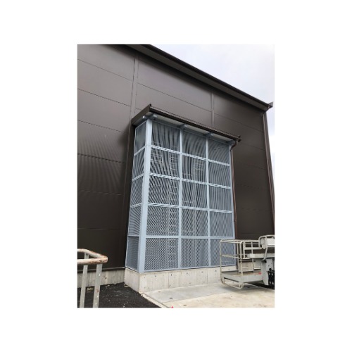 Metalowa konstrukcja osłonowa z kratką, przylegająca do brązowej ściany budynku. Widoczny fragment podnośnika koszowego.