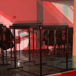 Szereg wieszaków na ubrania w szatni, oświetlonych czerwonym światłem, tworzących rytmiczny wzór. Widoczna metalowa siatka w tle.