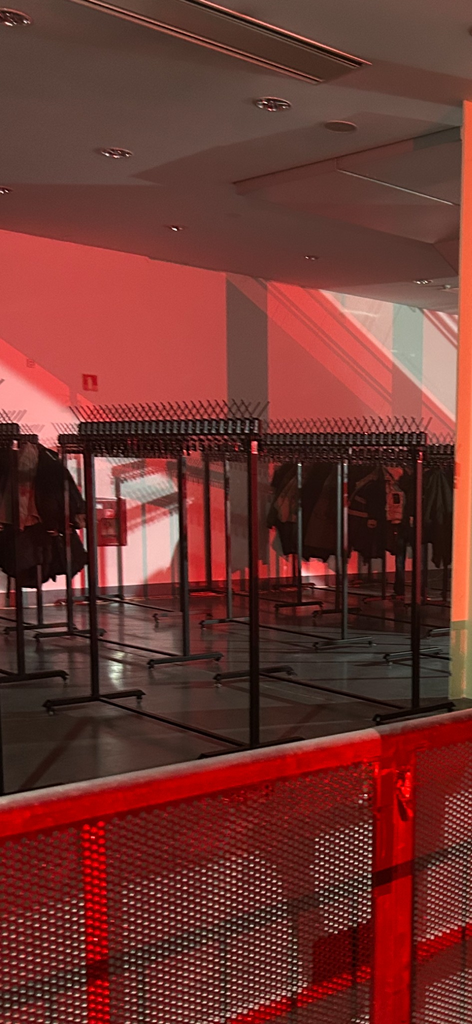 Szereg wieszaków na ubrania w szatni, oświetlonych czerwonym światłem, tworzących rytmiczny wzór. Widoczna metalowa siatka w tle.