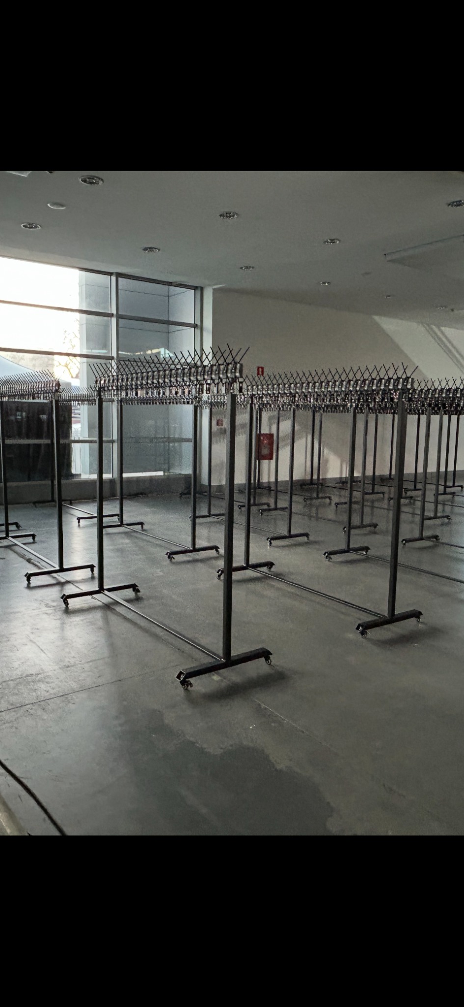Rzędy pustych, czarnych wieszaków na kółkach w jasnym wnętrzu, prawdopodobnie magazyn lub sala eventowa, przygotowane do użycia, widok z perspektywy.