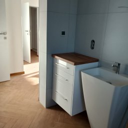 Artur Janura - Nowoczesna łazienka z białą szafką z drewnianym blatem i umywalką w kształcie kolumny. Parkiet w jodełkę. Minimalistyczny design, jasne wnętrze.