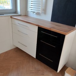 Artur Janura - Nowoczesna komoda łazienkowa z drewnianym blatem, fronty w kolorze białym i czarnym, metalowe uchwyty. Widoczny grzejnik drabinkowy i fragment okna.