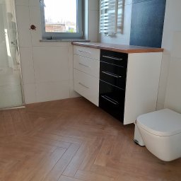 Artur Janura - Nowoczesna łazienka z szafką pod umywalkę w kolorze biało-czarnym, drewnianym blatem i podłogą w jodełkę. Widoczna toaleta i kabina prysznicowa.