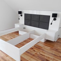 Artur Janura - Białe łóżko z wezgłowiem z szarymi panelami i lampkami nocnymi, w trakcie montażu, na drewnianej podłodze. Widoczne elementy konstrukcyjne i brak materaca.