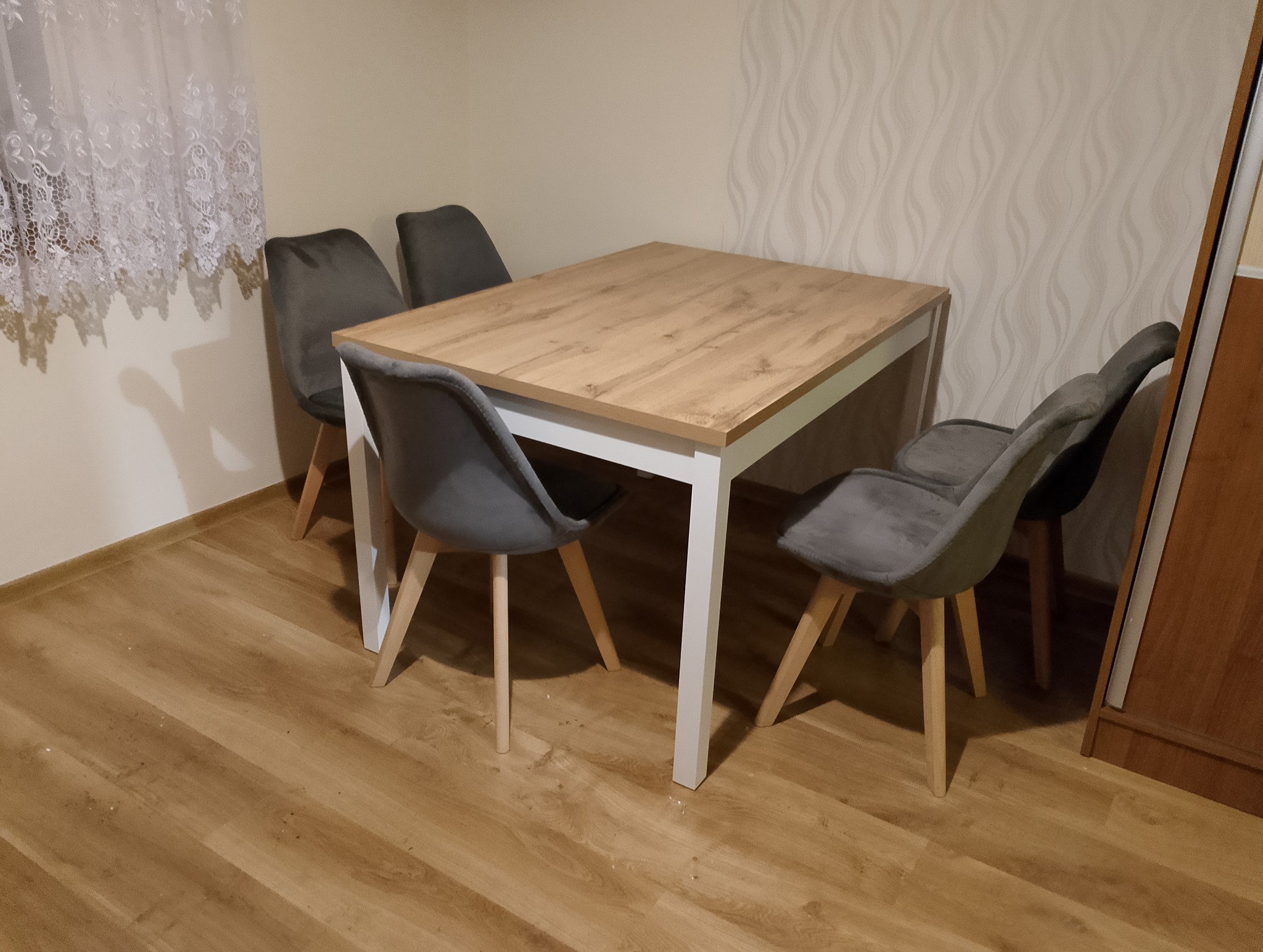 Drewniany stół z białymi nogami i szarymi krzesłami w pokoju z panelami podłogowymi i wzorzystą tapetą. Widok z perspektywy podłogi.