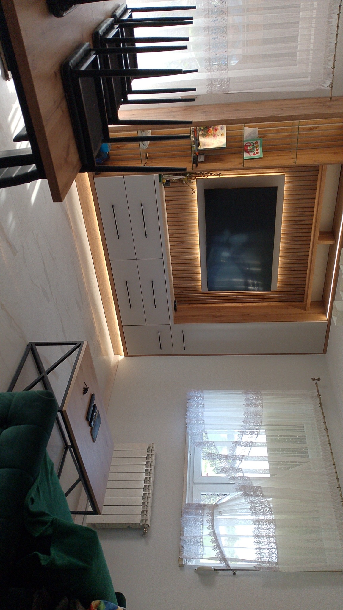 Nowoczesny salon z zabudową meblową pod telewizor, białe szafki z minimalistycznymi uchwytami, drewniane lamele i oświetlenie LED. Widok na okno z firankami.