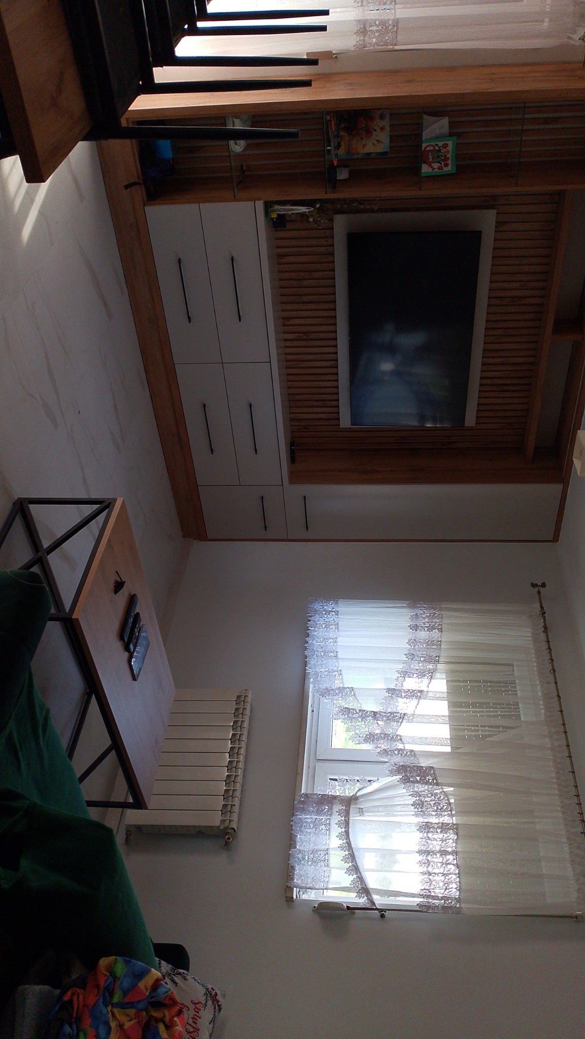 Nowoczesna zabudowa TV z szafkami i półkami w salonie w Sulmierzycach. Białe fronty szafek, drewniane wykończenie, minimalistyczny design. Widok na okno z firanką.