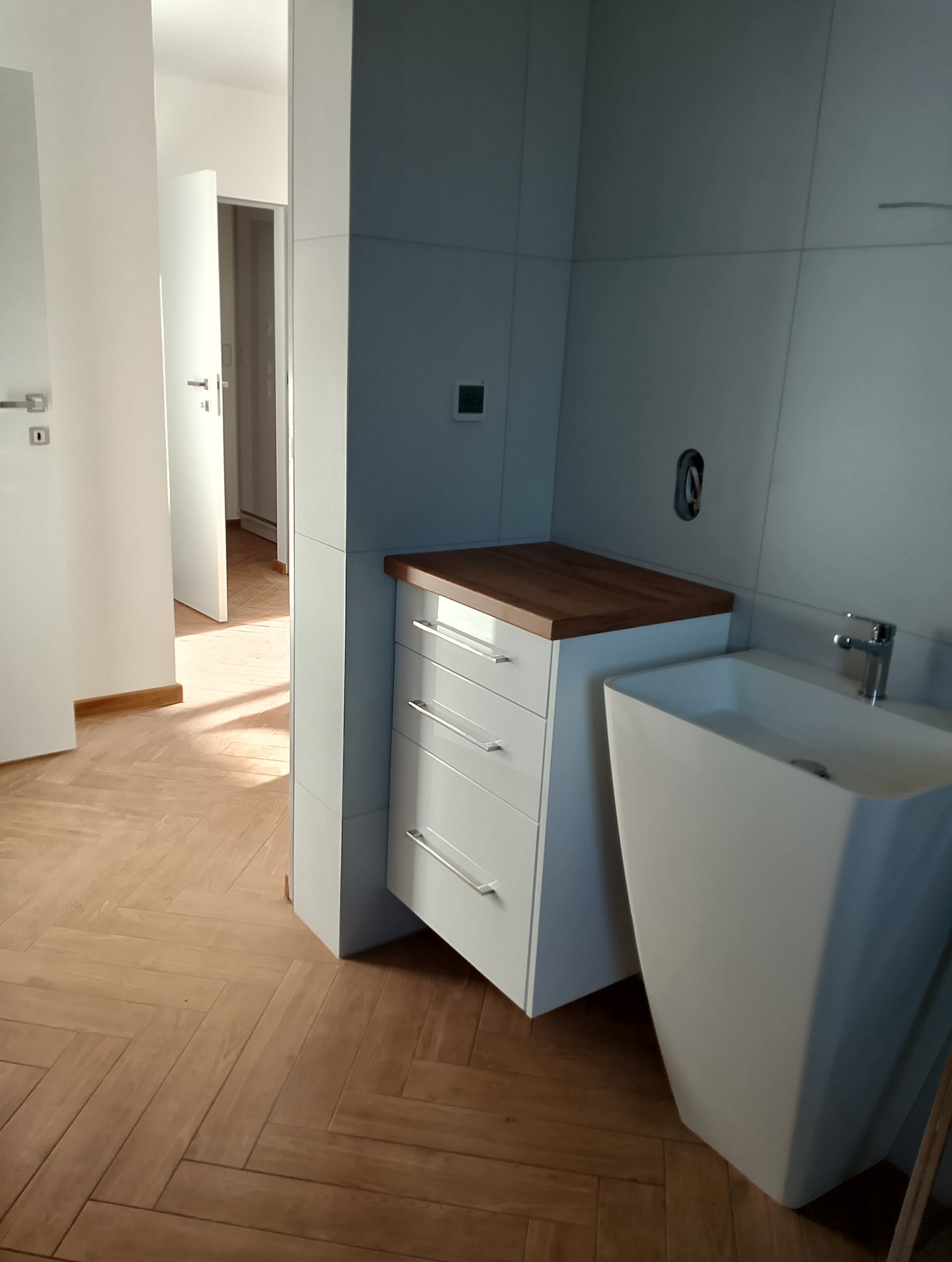 Nowoczesna łazienka z białą szafką z drewnianym blatem i umywalką w kształcie kolumny. Parkiet w jodełkę. Minimalistyczny design, jasne wnętrze.