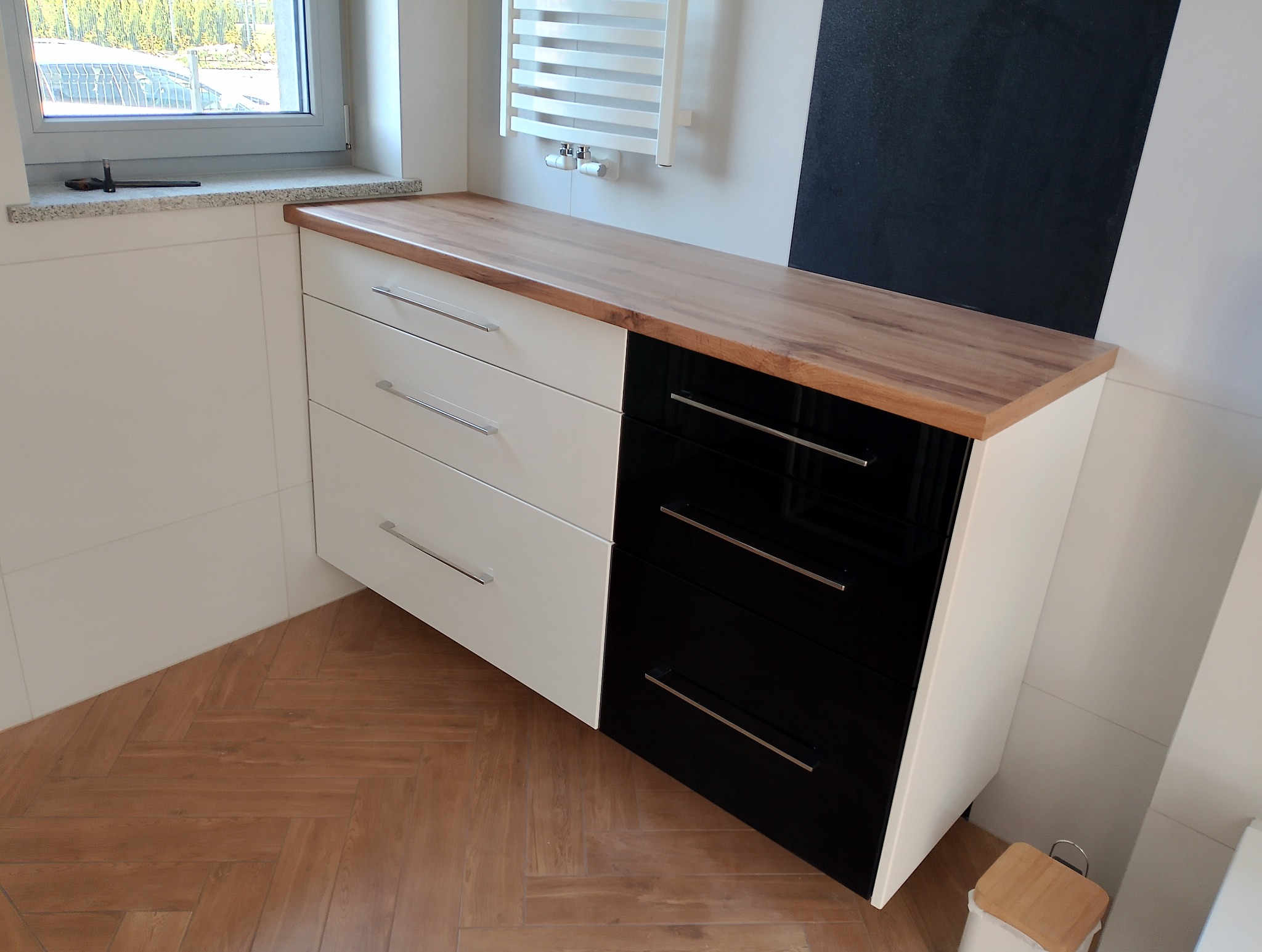 Nowoczesna komoda łazienkowa z drewnianym blatem, fronty w kolorze białym i czarnym, metalowe uchwyty. Widoczny grzejnik drabinkowy i fragment okna.