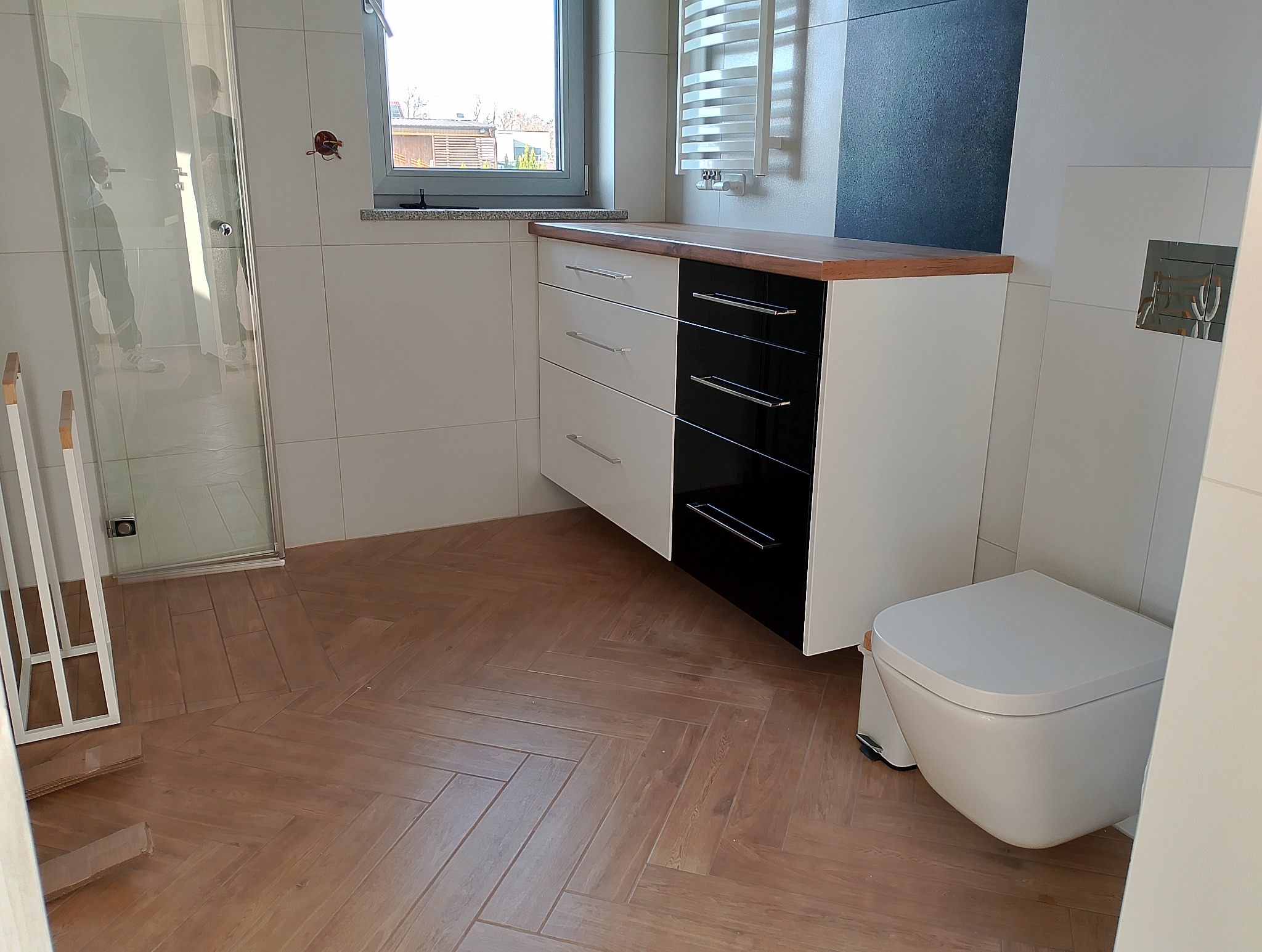 Nowoczesna łazienka z szafką pod umywalkę w kolorze biało-czarnym, drewnianym blatem i podłogą w jodełkę. Widoczna toaleta i kabina prysznicowa.