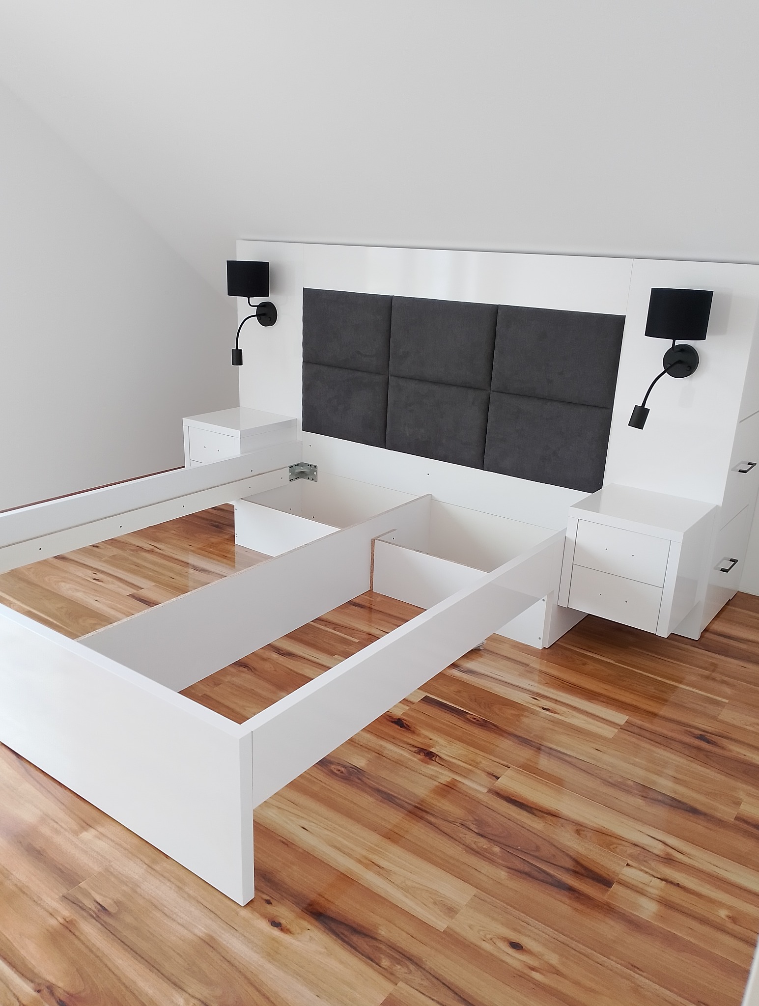Białe łóżko z wezgłowiem z szarymi panelami i lampkami nocnymi, w trakcie montażu, na drewnianej podłodze. Widoczne elementy konstrukcyjne i brak materaca.