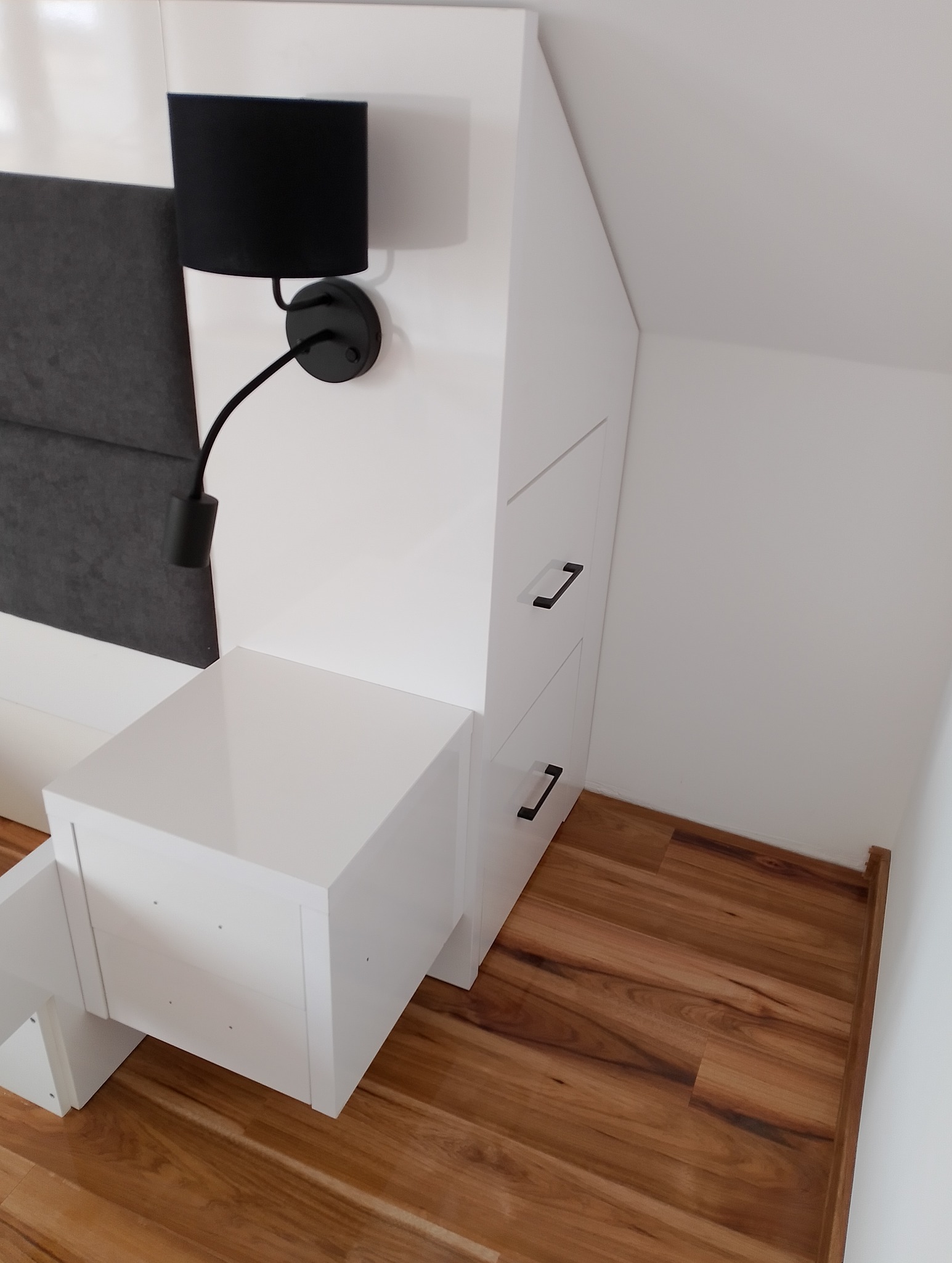 Biała szafka nocna z szufladami i czarną lampką na ścianie, dopasowana do skosu dachu. Drewniana podłoga i szary zagłówek łóżka w tle.