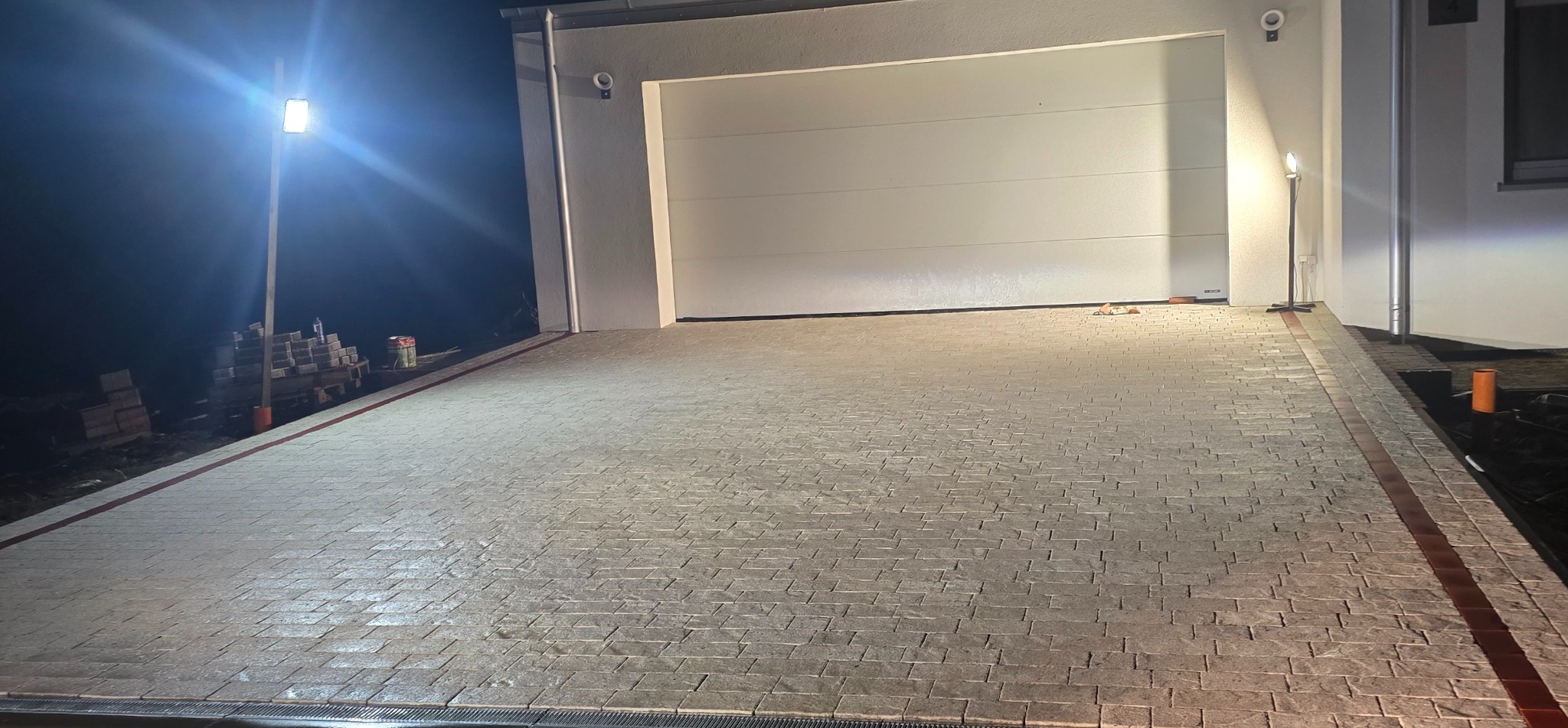 Ukończony podjazd z kostki brukowej w Chojnicach, oświetlony lampami. Widoczny garaż i fragment domu. Ciemne tło nocne, kostka w odcieniach szarości z bordową obwódką.