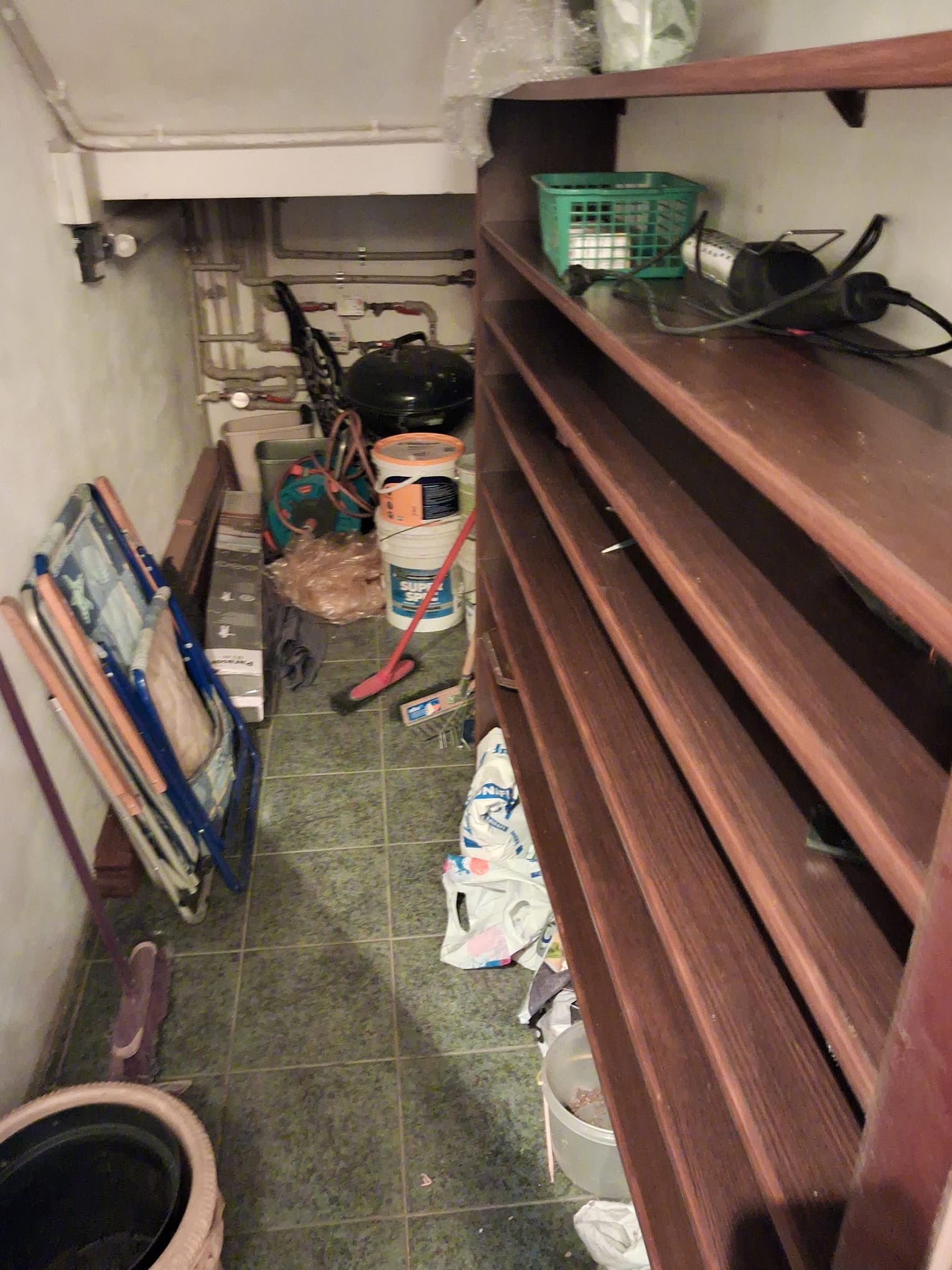 Zagracone pomieszczenie gospodarcze z regałem, wiadrami, sprzętem i różnymi przedmiotami. Widoczne rury i instalacje na ścianie. Podłoga wyłożona płytkami.