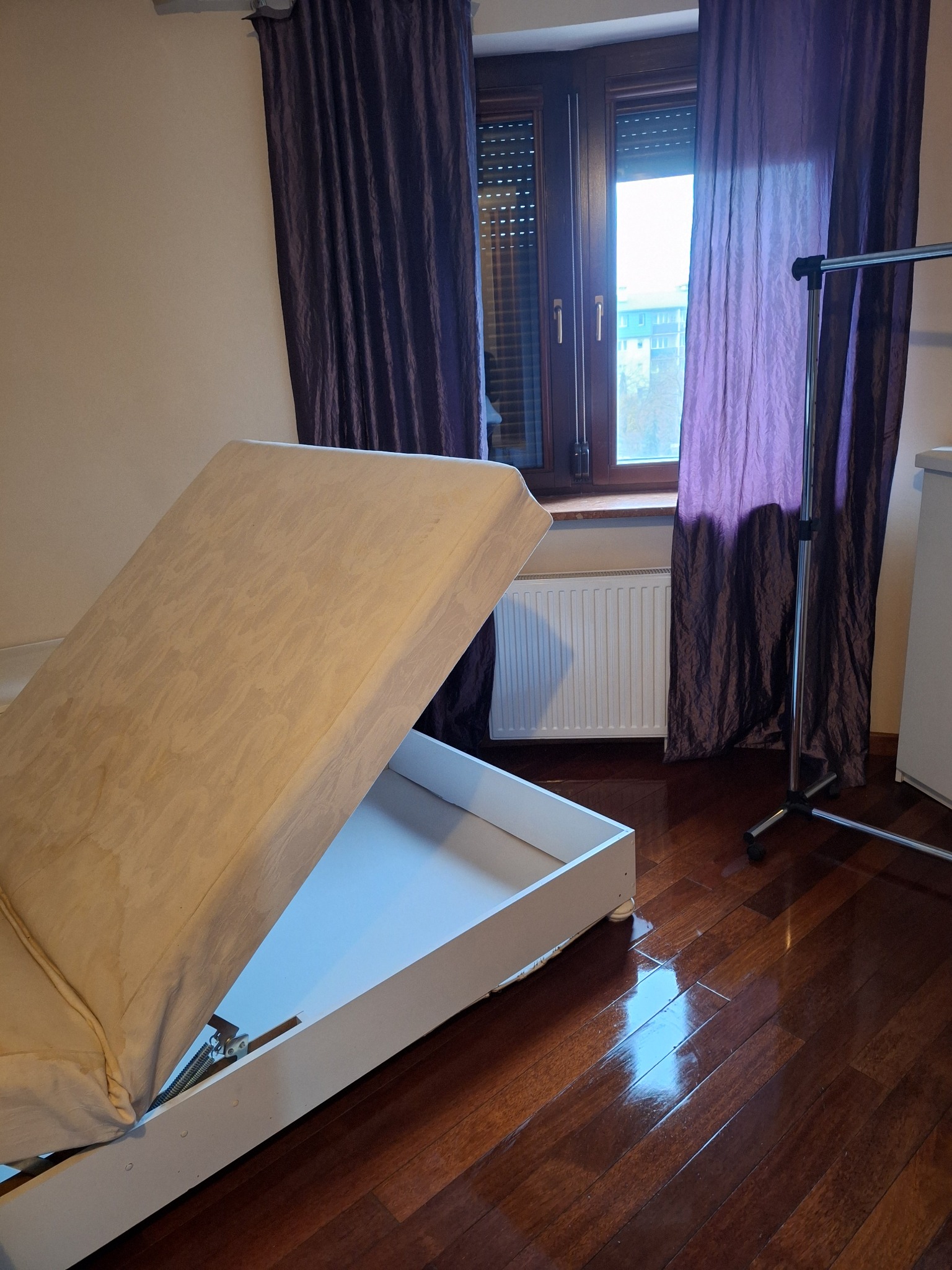 Otwarty tapczan z pojemnikiem na pościel na tle parkietu, okna z fioletowymi zasłonami i grzejnika. Widoczny wieszak na ubrania. Przygotowanie do sprzątania.