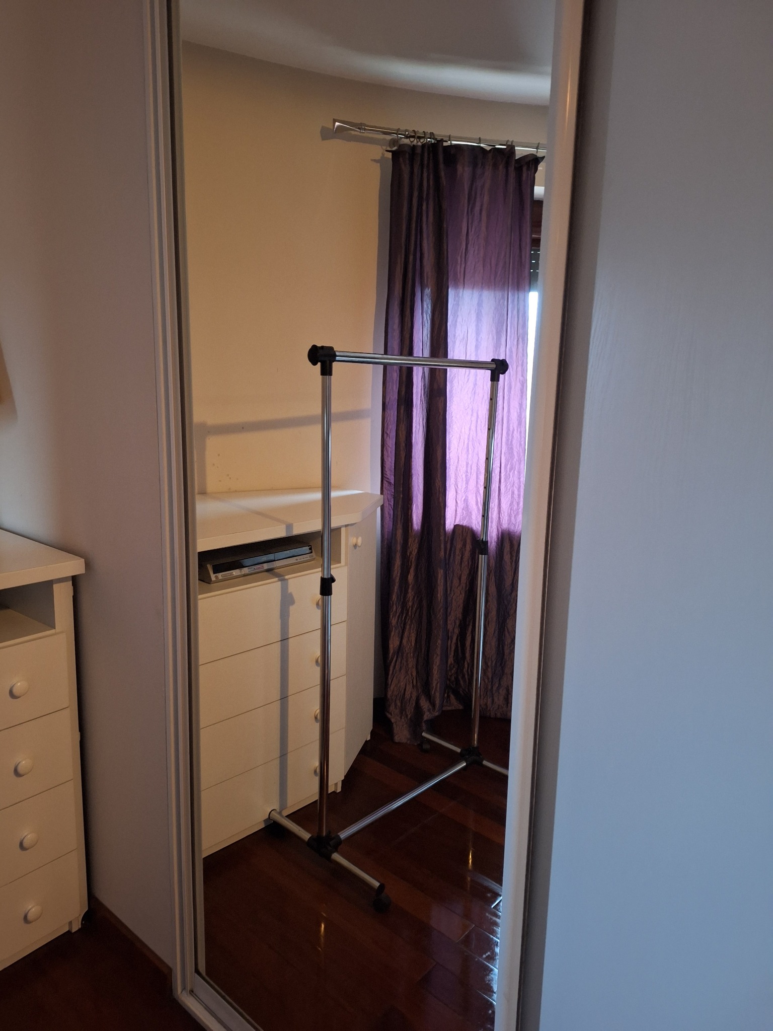 Odbicie w lustrze ukazuje uporządkowane wnętrze pokoju w Warszawie. Widoczna komoda, wieszak na ubrania i zasłony, co sugeruje dbałość o detale i czystość.