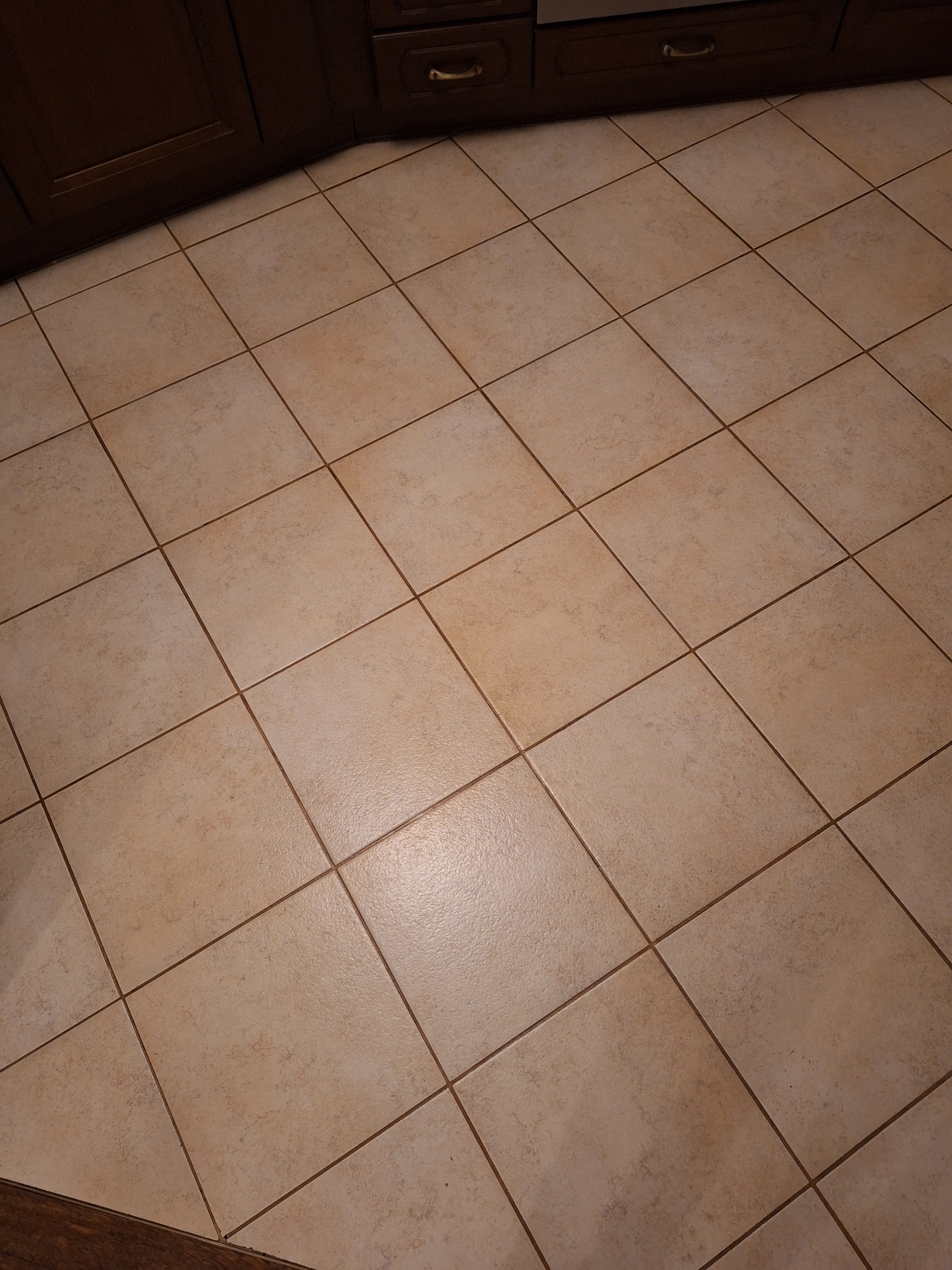 Beżowe, kwadratowe płytki podłogowe w kuchni, ułożone pod kątem, z widocznymi fugami i fragmentem ciemnobrązowej szafki kuchennej w rogu. Widok z góry.