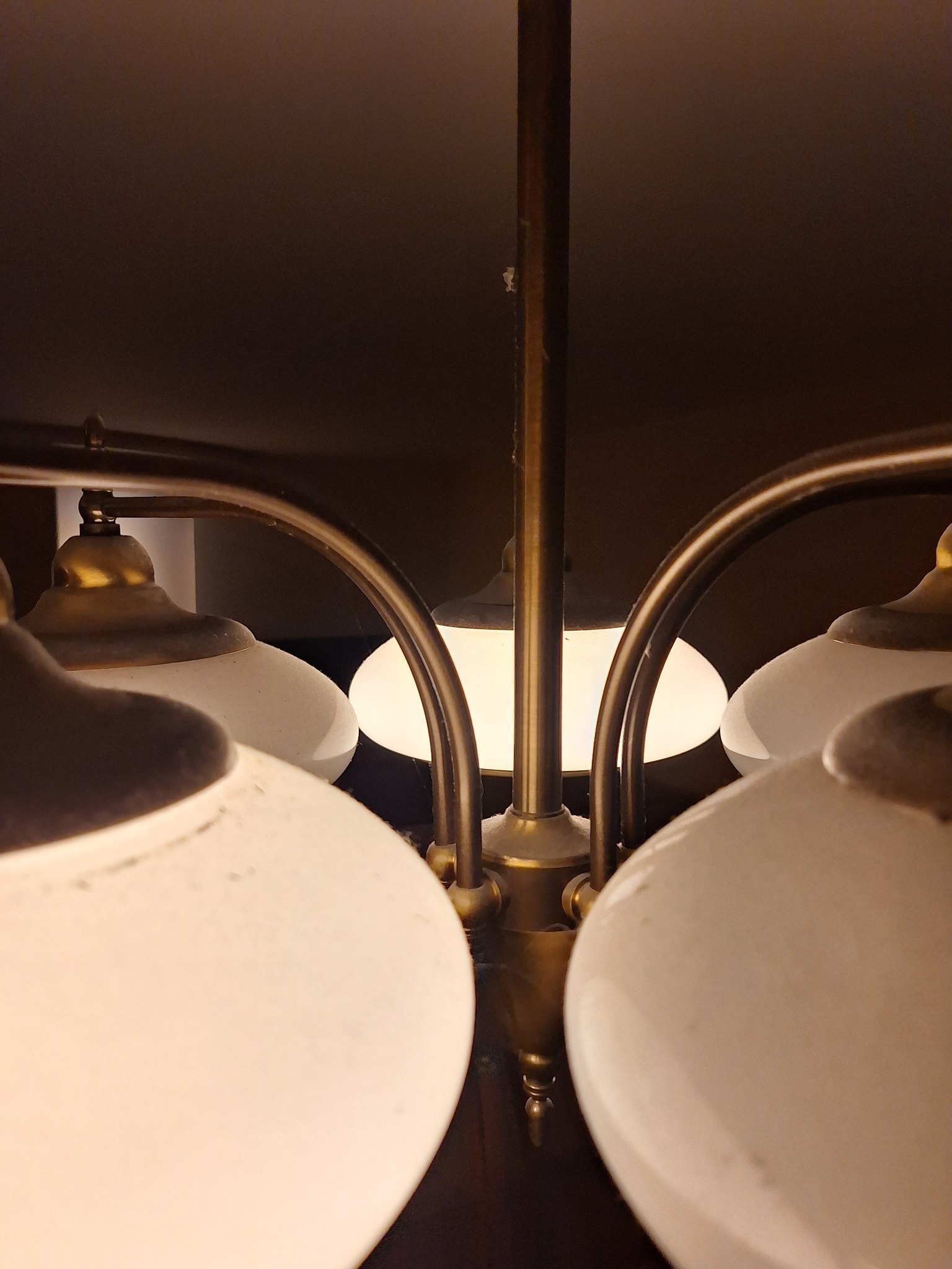 Mosiężny żyrandol z mlecznymi kloszami, widok z dołu, po czyszczeniu. Detal zabrudzeń na kloszach i kurzu na konstrukcji. Ciepłe światło żarówek.