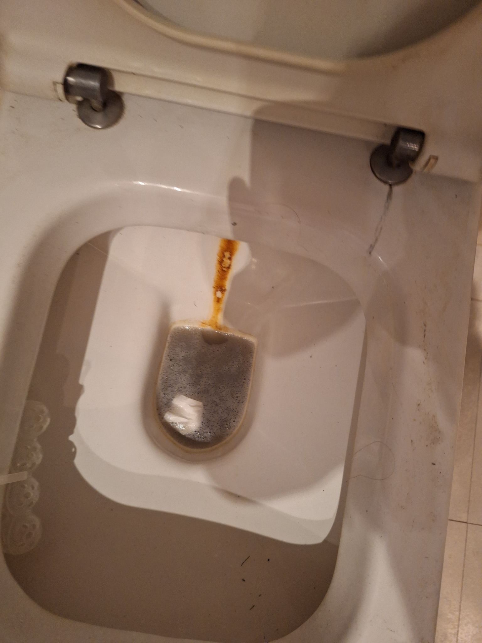 Z bliska: muszla klozetowa z osadem i środkiem czyszczącym w kształcie tabletki. Widoczne zabrudzenia na ceramice i ślady po twardej wodzie. Ujęcie z góry.