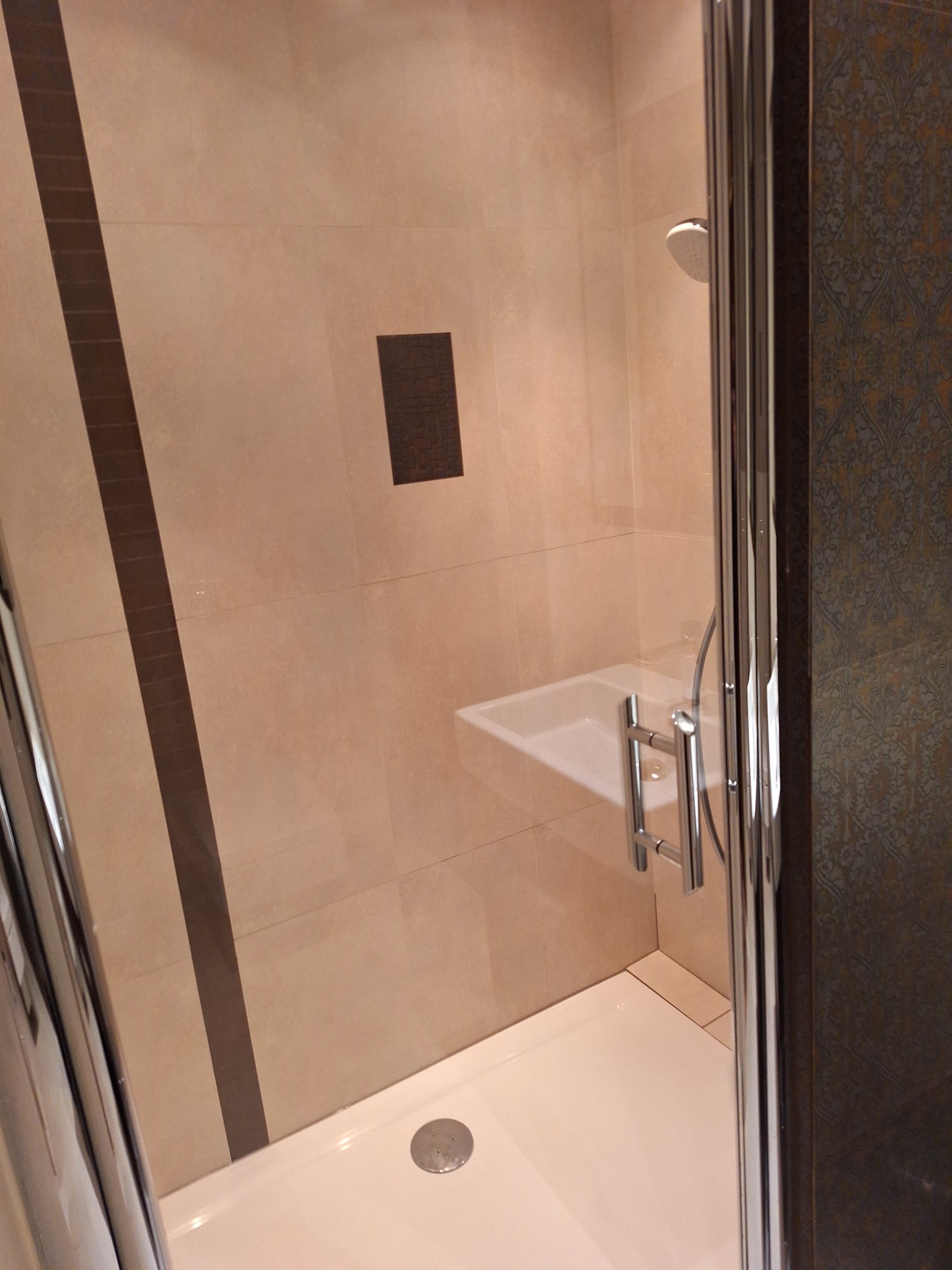 Czysta kabina prysznicowa z jasnymi płytkami i chromowanymi detalami. Widoczny brodzik, bateria i deszczownica. Minimalistyczny design, świeżo po sprzątaniu.