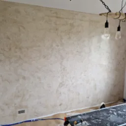 Ściana po nałożeniu dekoracyjnej, beżowej gładzi w stylu weneckim. Widoczne narzędzia na stole roboczym. Uwagę przykuwa lampa sufitowa z drewnianym elementem.