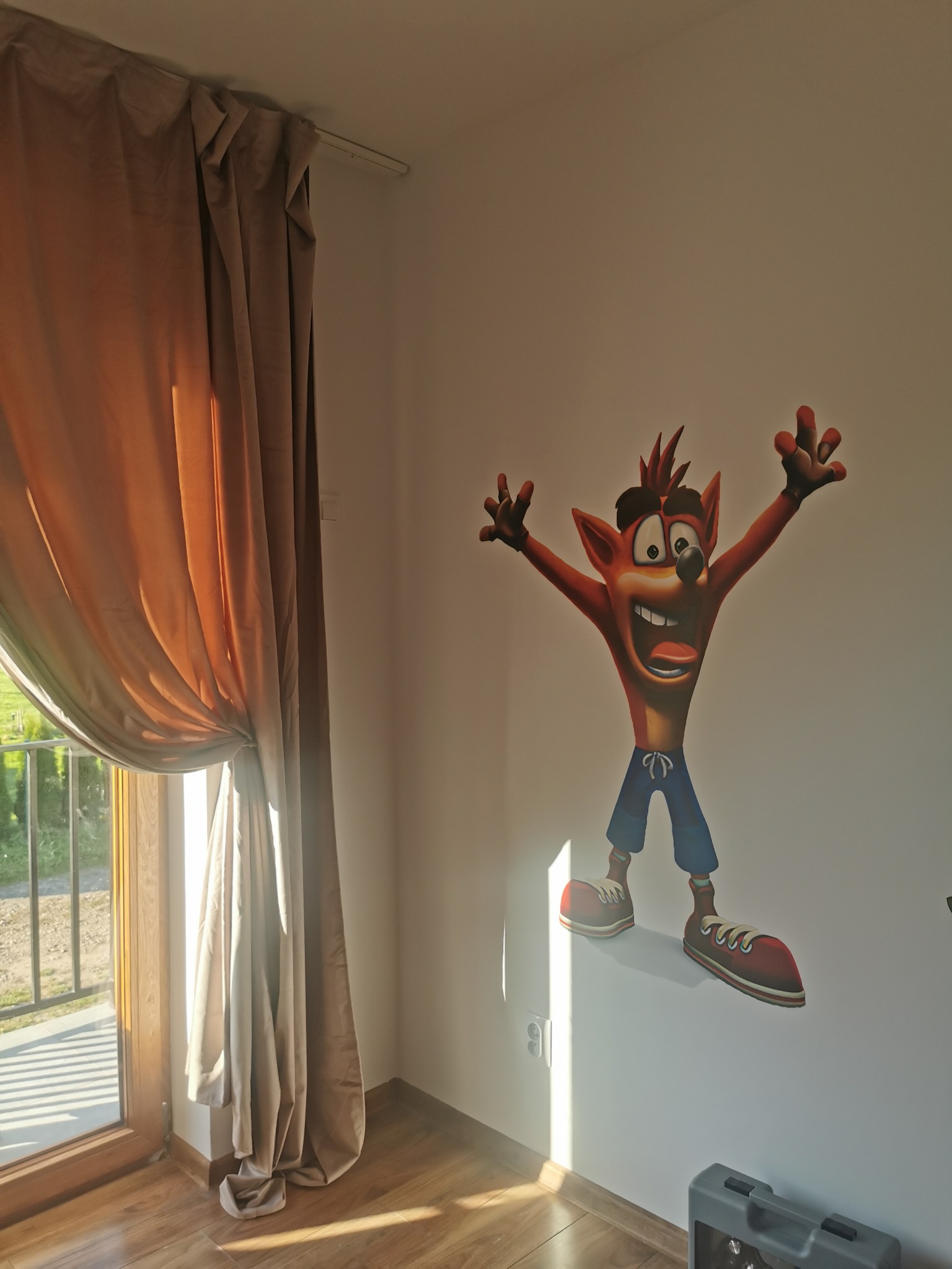 Pokój z naklejką na ścianie przedstawiającą postać z gry Crash Bandicoot, obok okno z zasłoną w odcieniach brązu. Na podłodze widoczny fragment skrzynki narzędziowej.