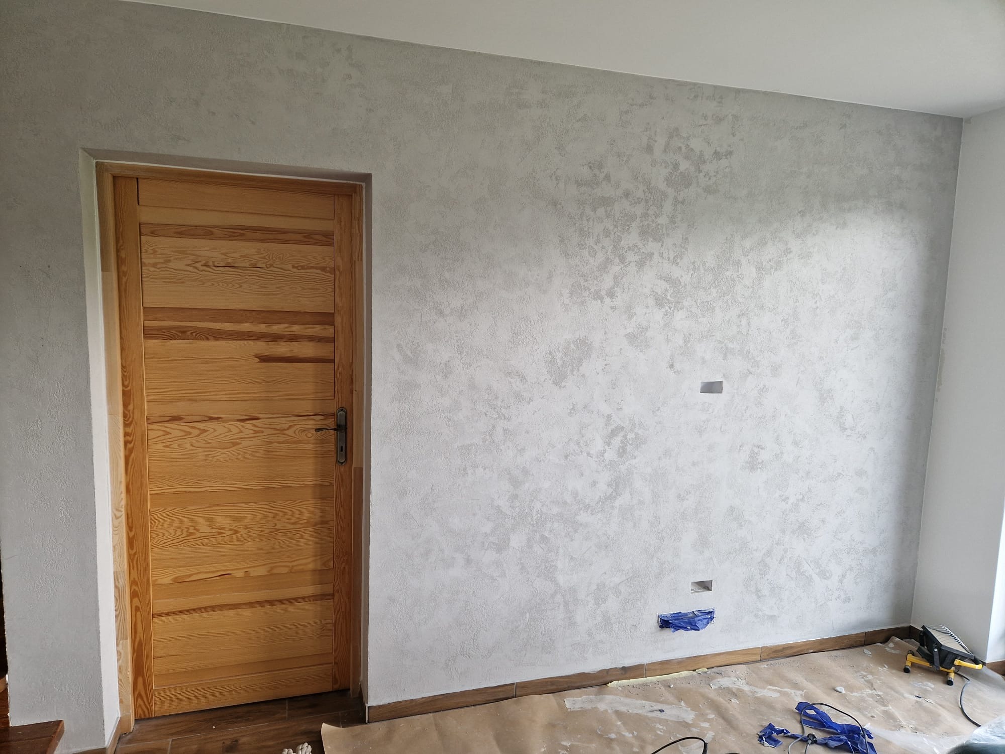 Ściana z efektem dekoracyjnym w odcieniach szarości, drewniane drzwi po lewej stronie, podłoga zabezpieczona folią malarską, widoczne gniazdka elektryczne.