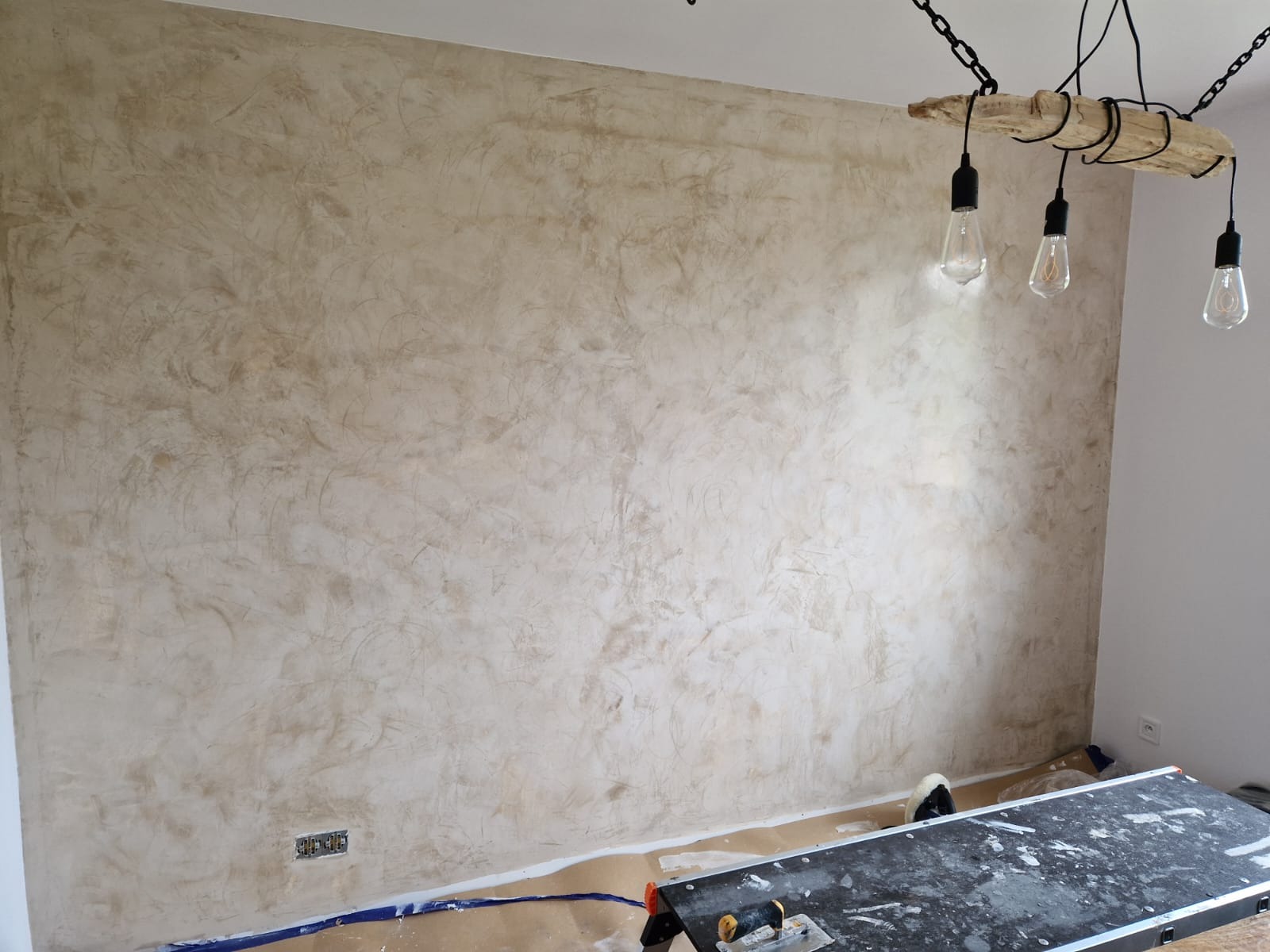 Ściana po nałożeniu dekoracyjnej, beżowej gładzi w stylu weneckim. Widoczne narzędzia na stole roboczym. Uwagę przykuwa lampa sufitowa z drewnianym elementem.