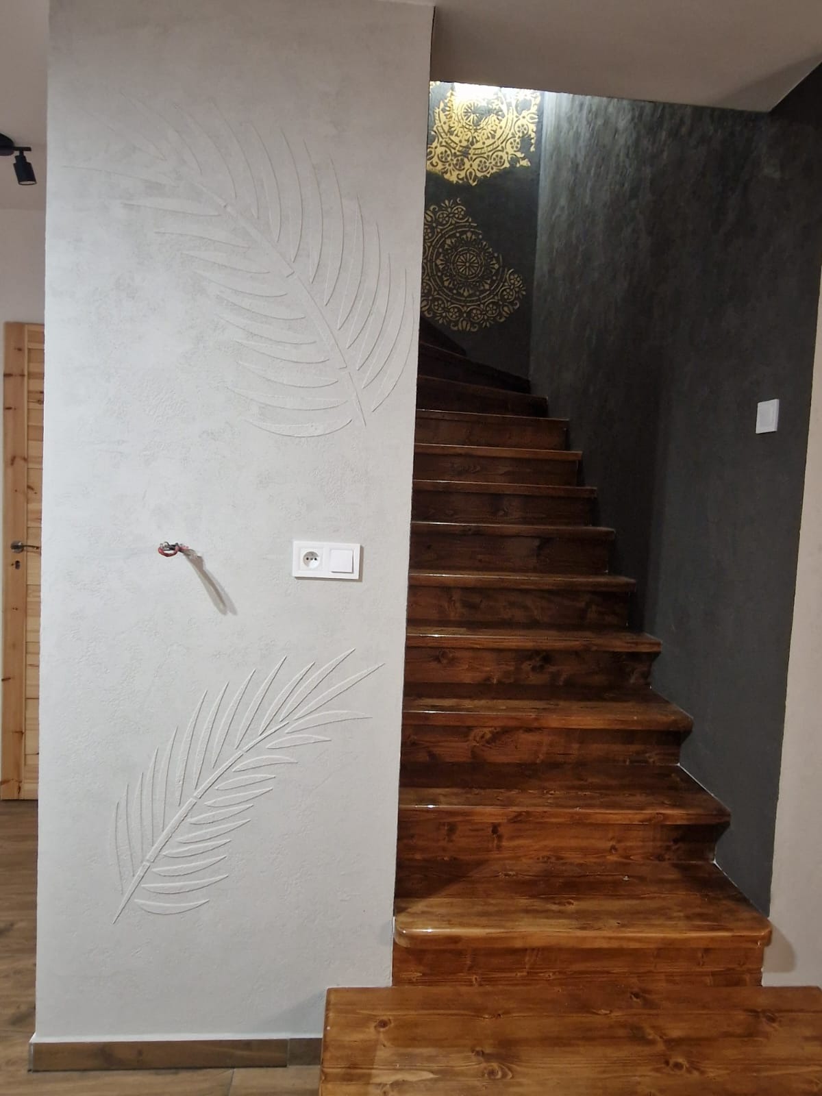 Wykończenie ściany przy schodach z dekoracyjnym, wypukłym motywem liści. Schody drewniane, lakierowane. Fragment instalacji elektrycznej. Ciemna ściana z ornamentem.