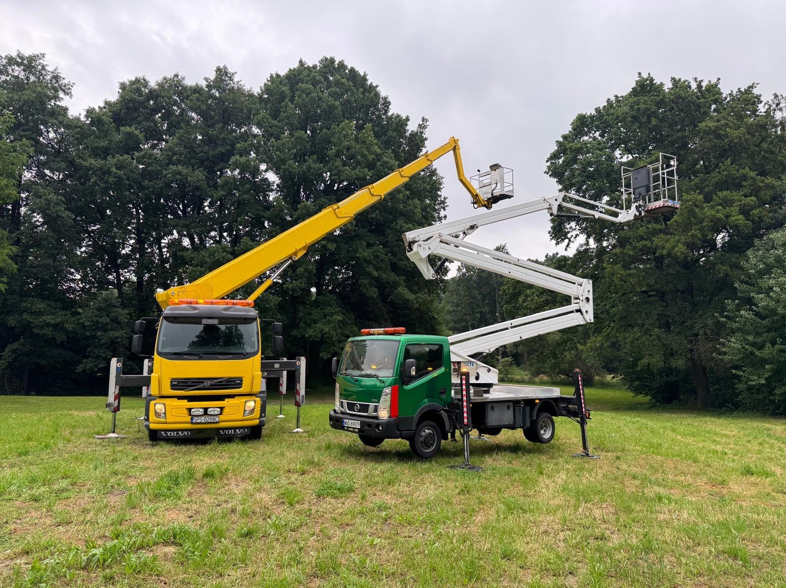 Dwa podnośniki koszowe, żółty Volvo i zielony Nissan, na trawiastym terenie w Żorach. Kosze wysunięte, gotowe do pracy przy drzewach. Pochmurne niebo w tle.