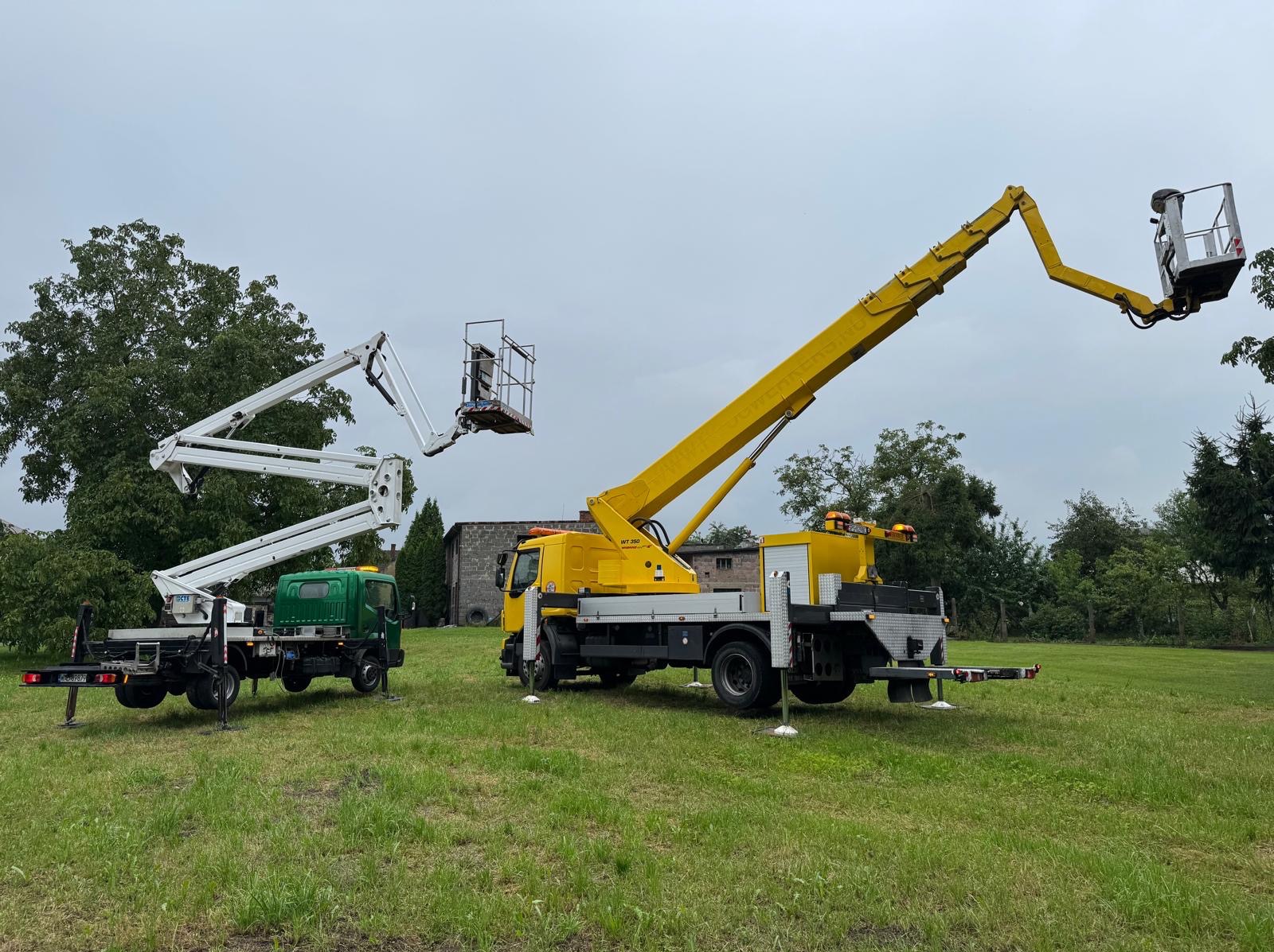 Dwa podnośniki koszowe: żółty i zielony, stoją na trawiastym terenie. Żółty podnośnik z wysuniętym ramieniem, biały podnośnik z koszem w górze. Pochmurne niebo w tle.