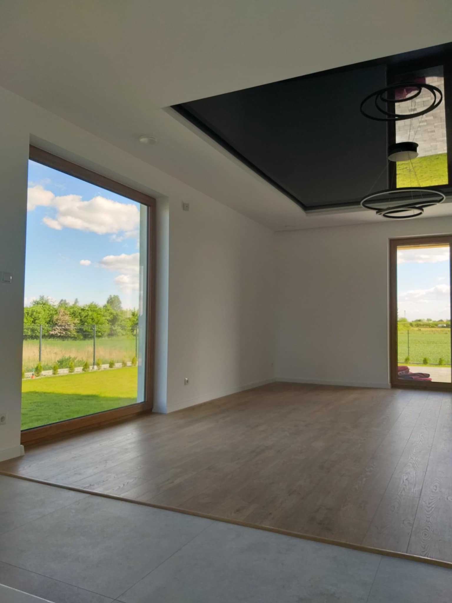 Jasne wnętrze z dużymi oknami i drewnianą podłogą. Na suficie czarna wnęka z nowoczesnym oświetleniem. Widok na zewnątrz z zielonym krajobrazem.