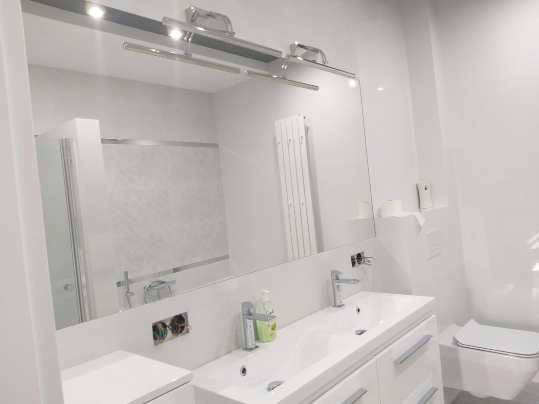 Nowoczesna, jasna łazienka z dużym lustrem, podwójną umywalką i toaletą w minimalistycznym stylu. Widoczne niedokończone podłączenia elektryczne.