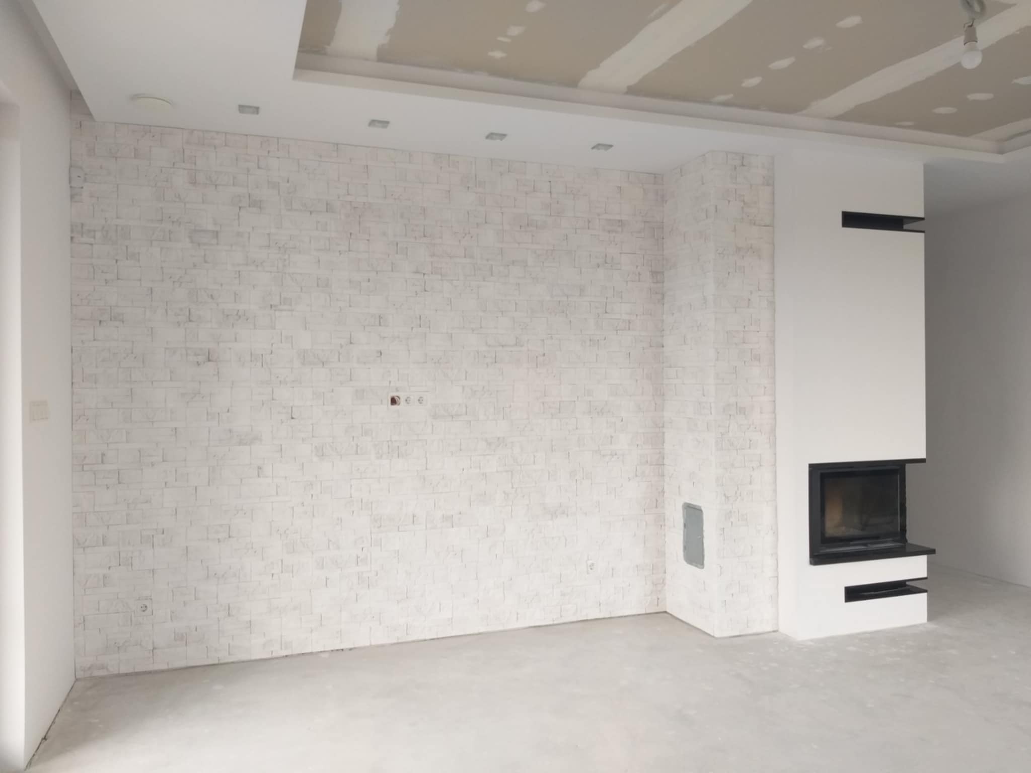 Ściana z białej cegły dekoracyjnej, kominek z czarną obudową, sufit z płyt karton-gips, szpachlowane łączenia, widok na wnętrze w trakcie wykańczania.