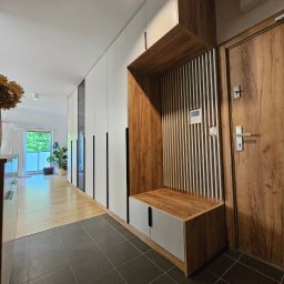 Manek Meble Kacper Manuszewski - Nowoczesna zabudowa przedpokoju z szafami wnękowymi w bieli i drewnie, z siedziskiem i dekoracyjnymi lamelami. Minimalistyczny design, czarne uchwyty, szara podłoga.