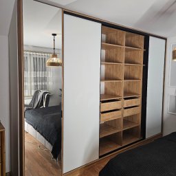 Manek Meble Kacper Manuszewski - Szafa wnękowa z przesuwnymi drzwiami: lustrzane, białe i drewniane panele. Wnętrze z półkami i szufladami. Widok na sypialnię w lustrze.