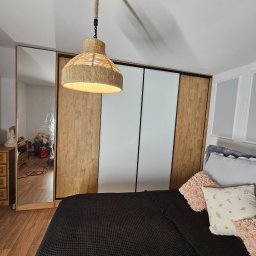 Manek Meble Kacper Manuszewski - Szafa wnękowa z przesuwnymi drzwiami: drewniane panele po bokach, białe w środku. W tle widoczne łóżko i lampa z abażurem z naturalnego sznurka.