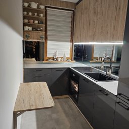 Manek Meble Kacper Manuszewski - Nowoczesna kuchnia z szarymi szafkami, drewnianymi akcentami i rozkładanym blatem. Widoczne półki z dekoracjami i odbicie w lustrzanych panelach nad blatem.