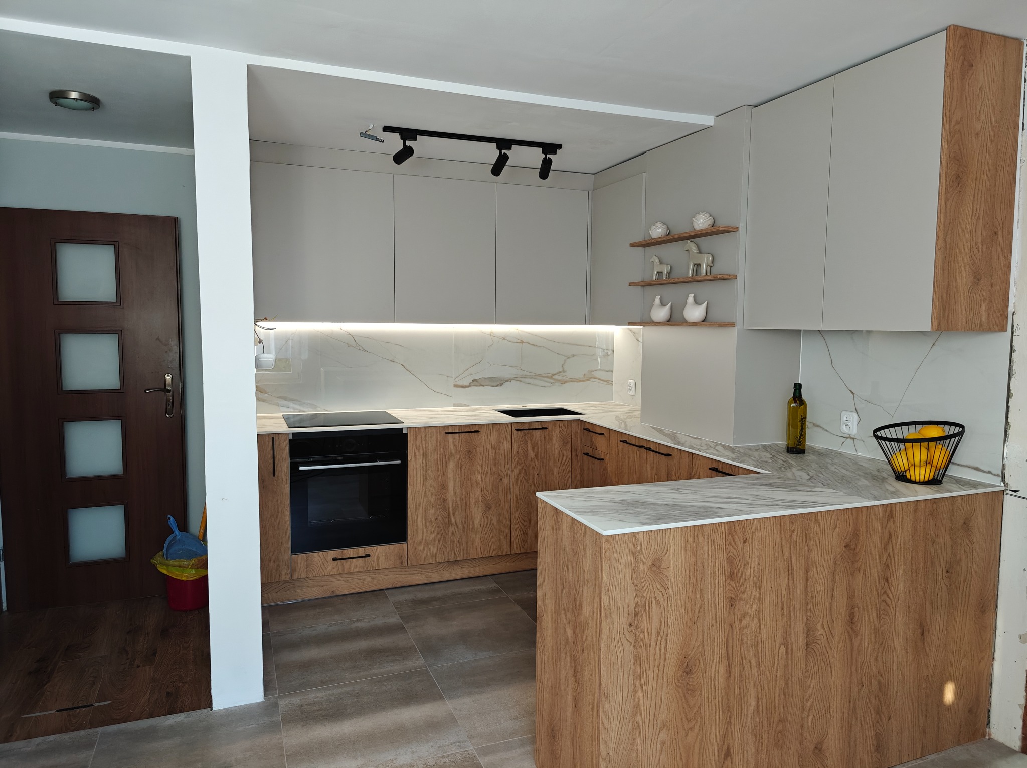 Nowoczesna kuchnia z drewnianymi i szarymi frontami, marmurowym blatem i oświetleniem punktowym. Na półkach dekoracje, a na blacie koszyk z cytrynami.