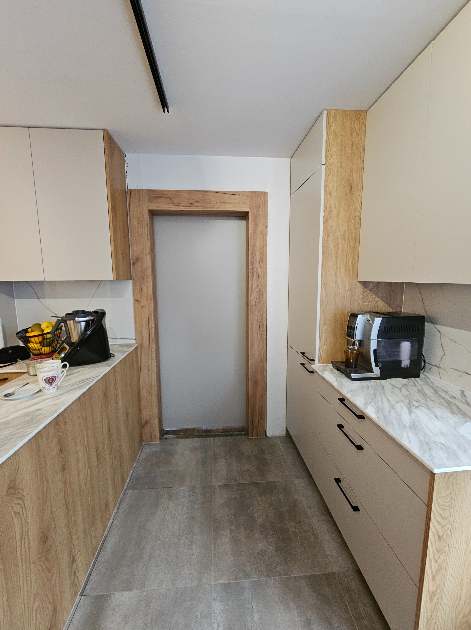 Nowoczesna kuchnia z drewnianymi akcentami: jasne szafki, marmurowy blat, ekspres do kawy i drzwi z drewnianą ramą. Minimalistyczny design i funkcjonalność.