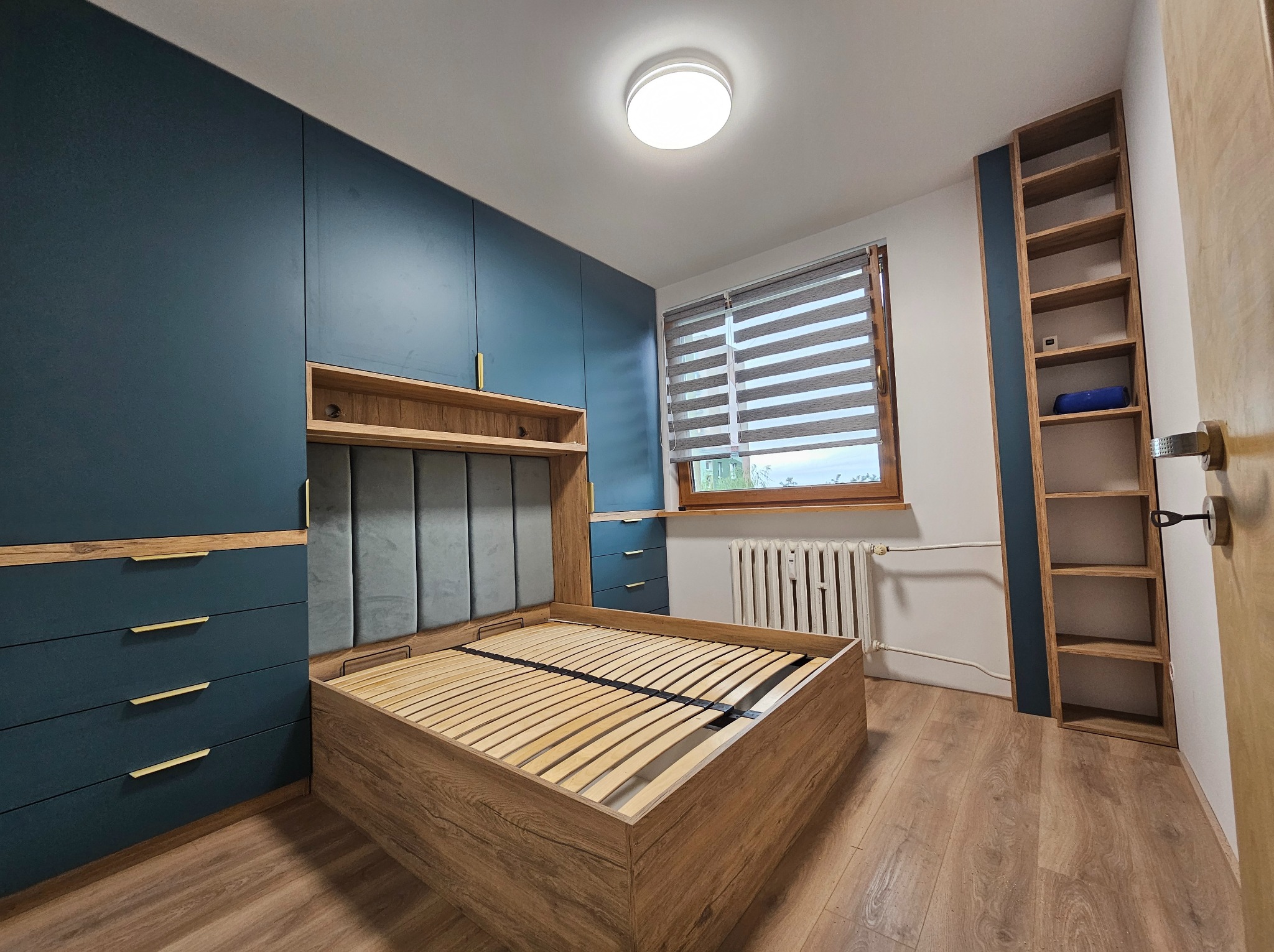 Kompletna sypialnia z łóżkiem z drewnianą ramą i wezgłowiem z szarej tapicerki, szafki nocne i szafa w zabudowie w kolorze granatowym z drewnianymi akcentami.